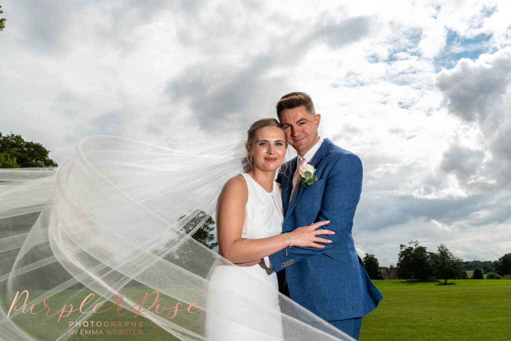 Bride and groom stood in front of stormy sky with brides veil swirling around them