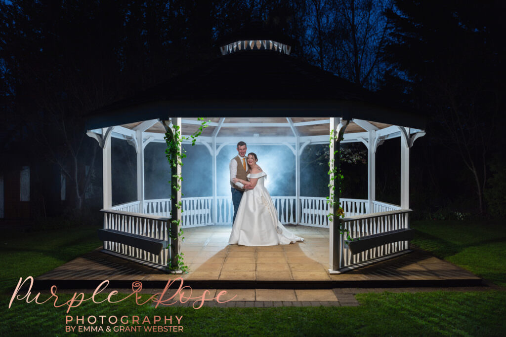 Night time photo of a bride and groom stood under a pergala surrounded by smoke on their wedding day in Milton Keynes