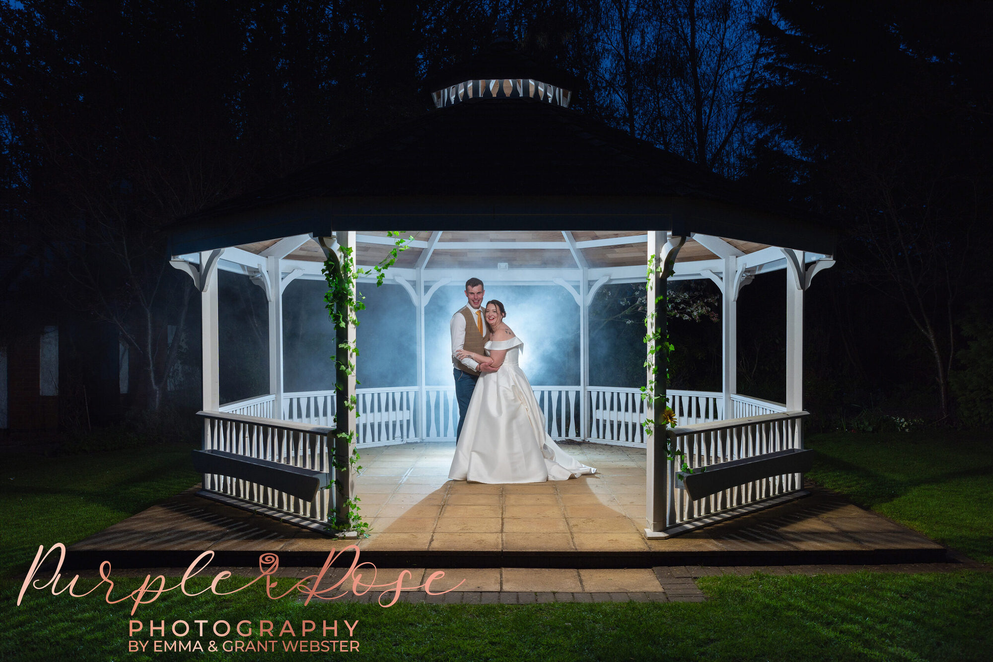 Night time photo of a bride and groom stood under a pergala surrounded by smoke on their wedding day in Milton Keynes