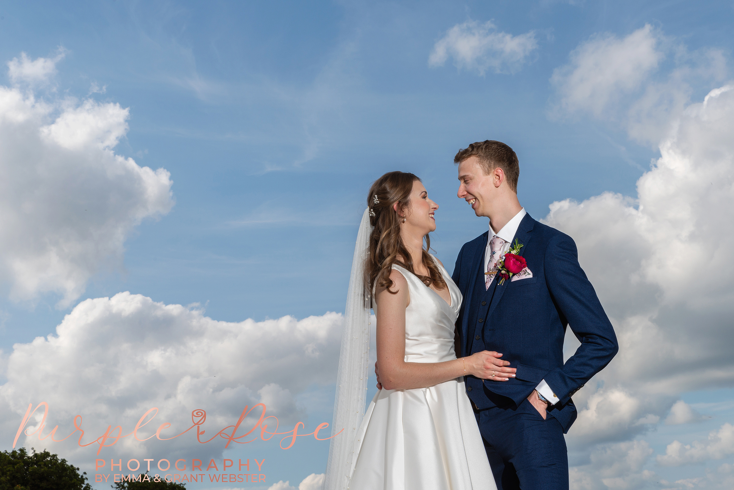 Photo of bride and groom smiling at each other in front of a blue sky on their wedding day in Milton Keynes