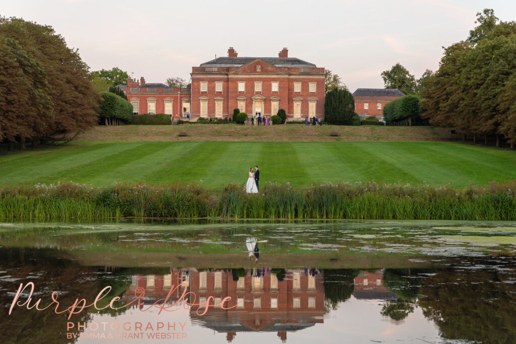 Photo of bride and groom from across a lake with their wedding venue behind them in Milton Keynes