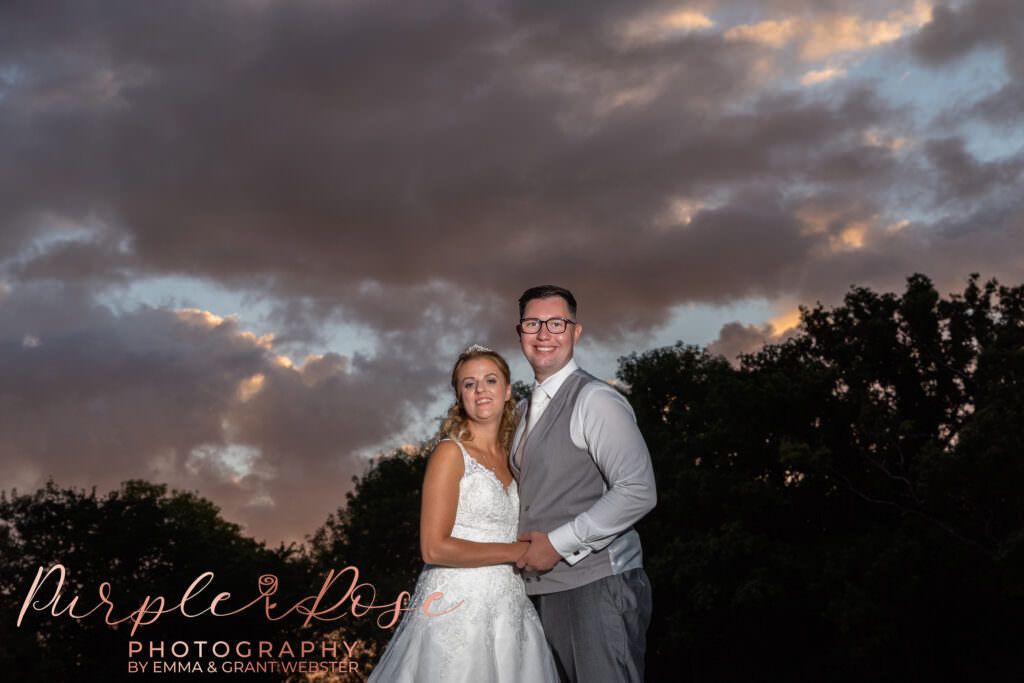 Bride and groom stood in front of a cloud filled sunset.