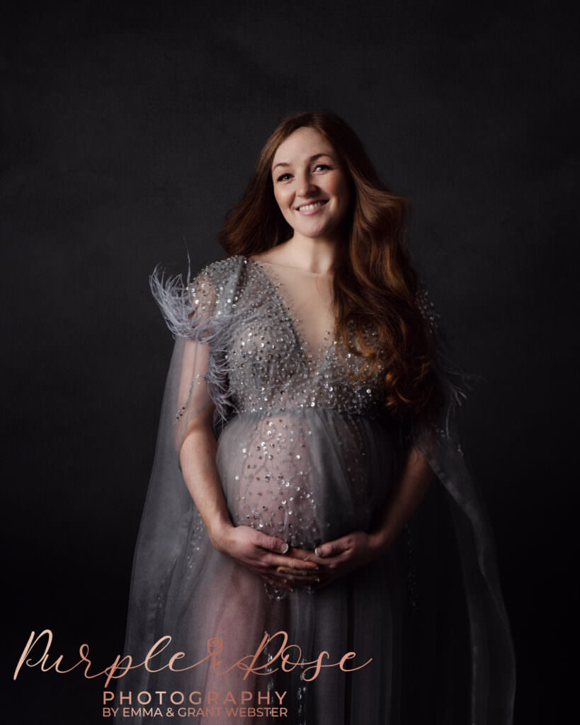 A pregnant woman in a grey dress smiling at the camera at her maternity photoshoot in Milton Keynes