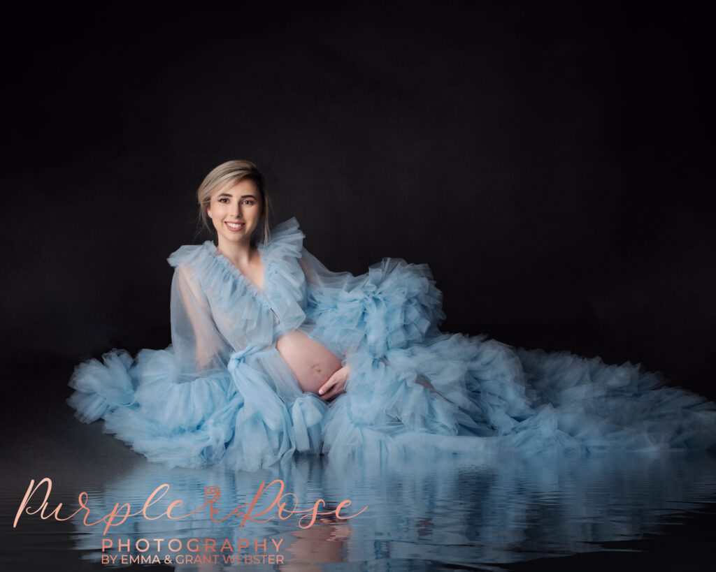 An expectant mother in pale blue tulle dress, laying on water like floor in a studio photo shoot in Milton Keynes