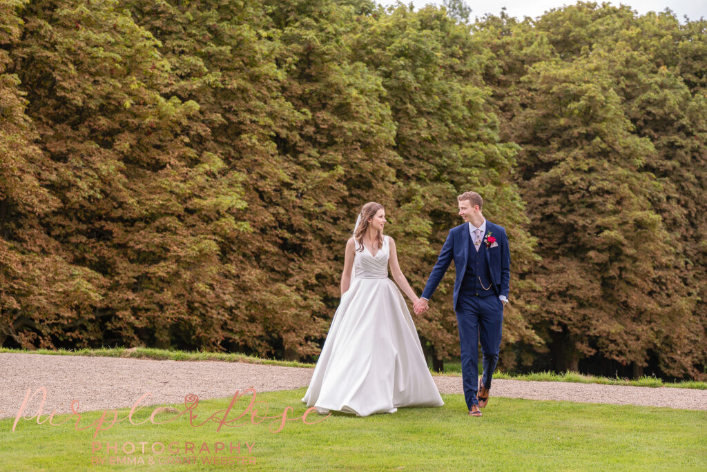 Photo of bride and groom walking hand in hand in front of autumn trees in Milton Keynes
