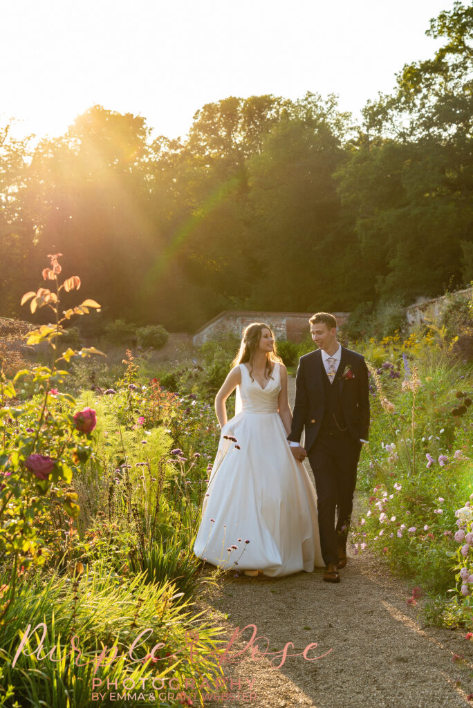 Photo of bride and groom walking hand in hand on a garden backlit at sunset on their wedding day in Milton Keynes