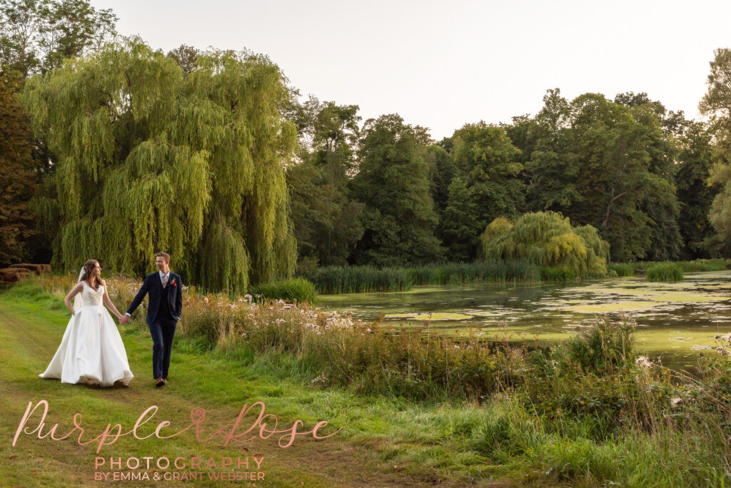 Photo of a bride and groom walking hand in hand by a lake on their wedding day.