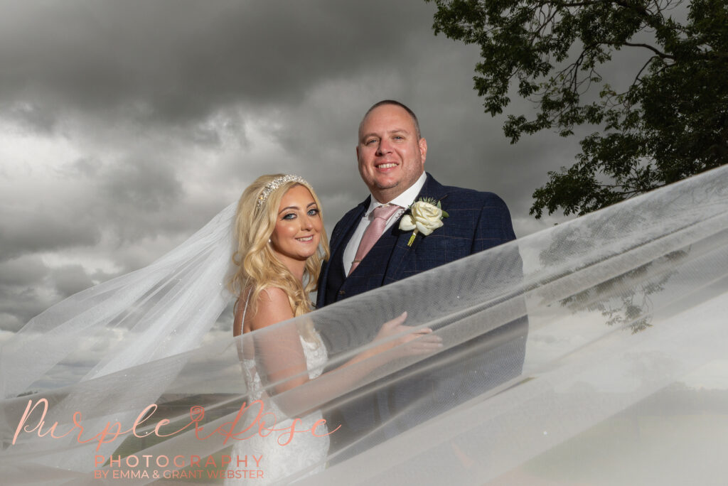 Photo of bride and groom stood in front of a stormy sky with thei bride veil swirling around them on their wedding day in Northampton
