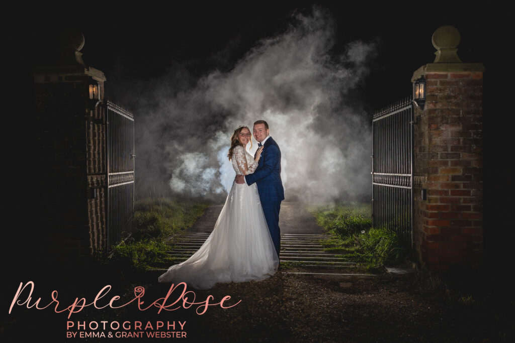 Photo of abride and groom at night in fornt of metal gates with smoke behind them on their wedding day in Milton Keynes